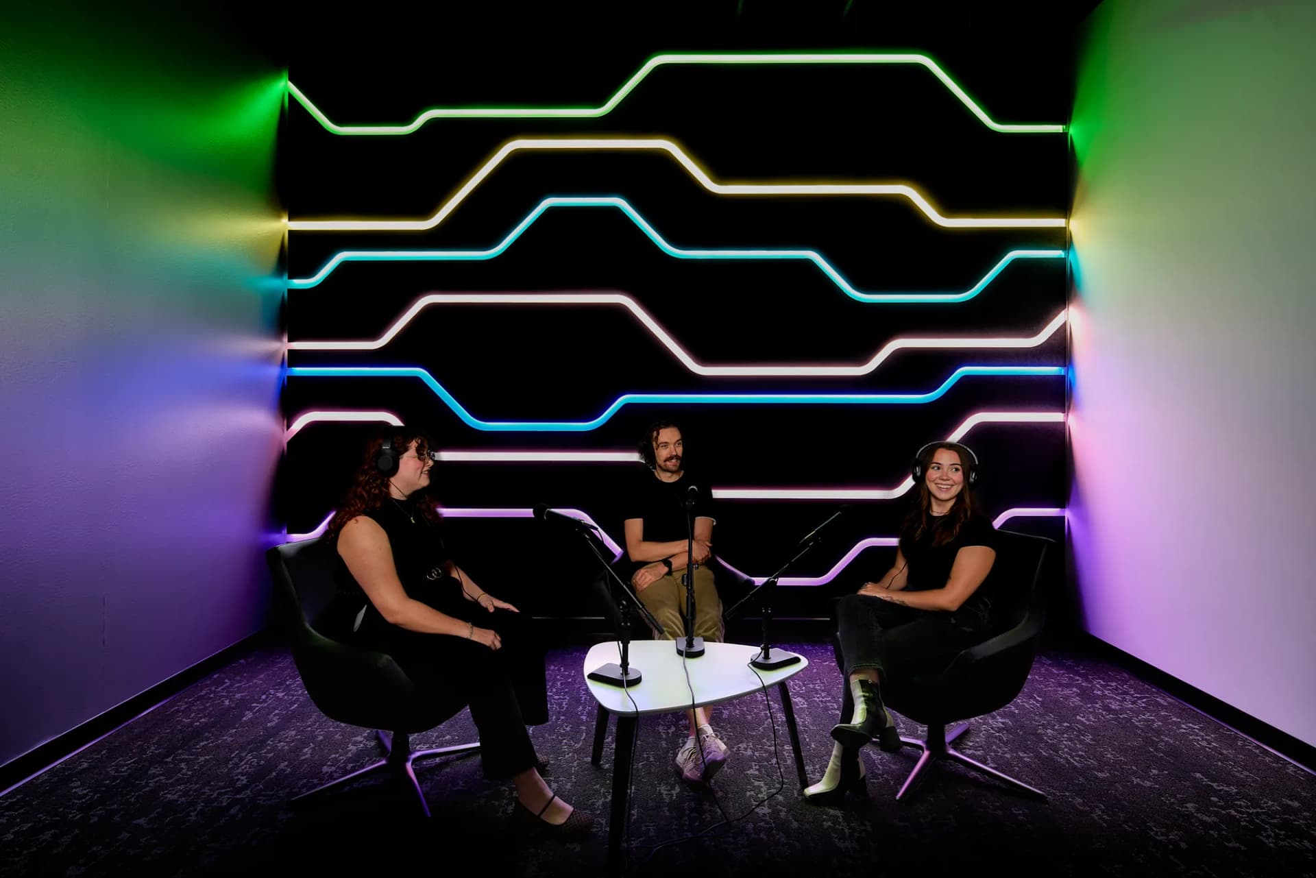 Podcast room with neon light lines across a black wall and three people seated in conversation